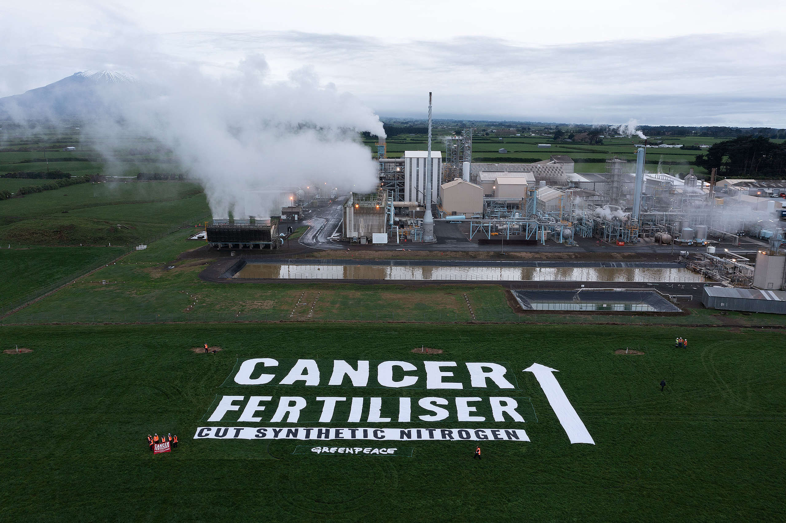 A factory with clouds of smoke has giant banner message in front saying 'cancer fertiliser, no synthetic nitrogen fertiliser'
