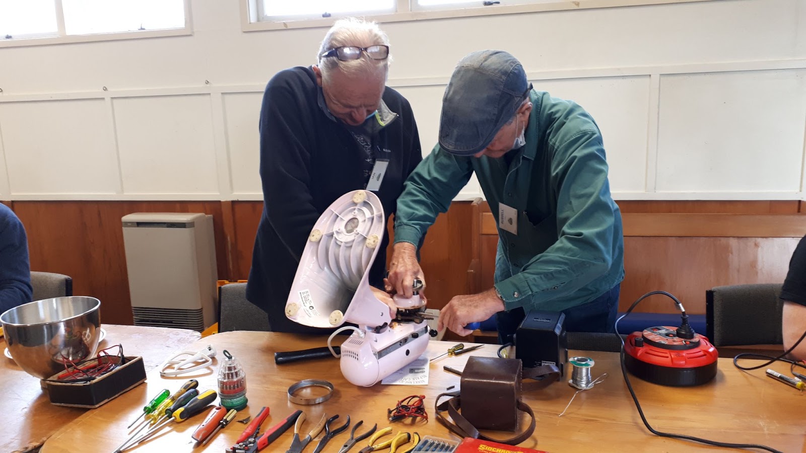 Two men are working at fixing a fan, on a table with many small tools