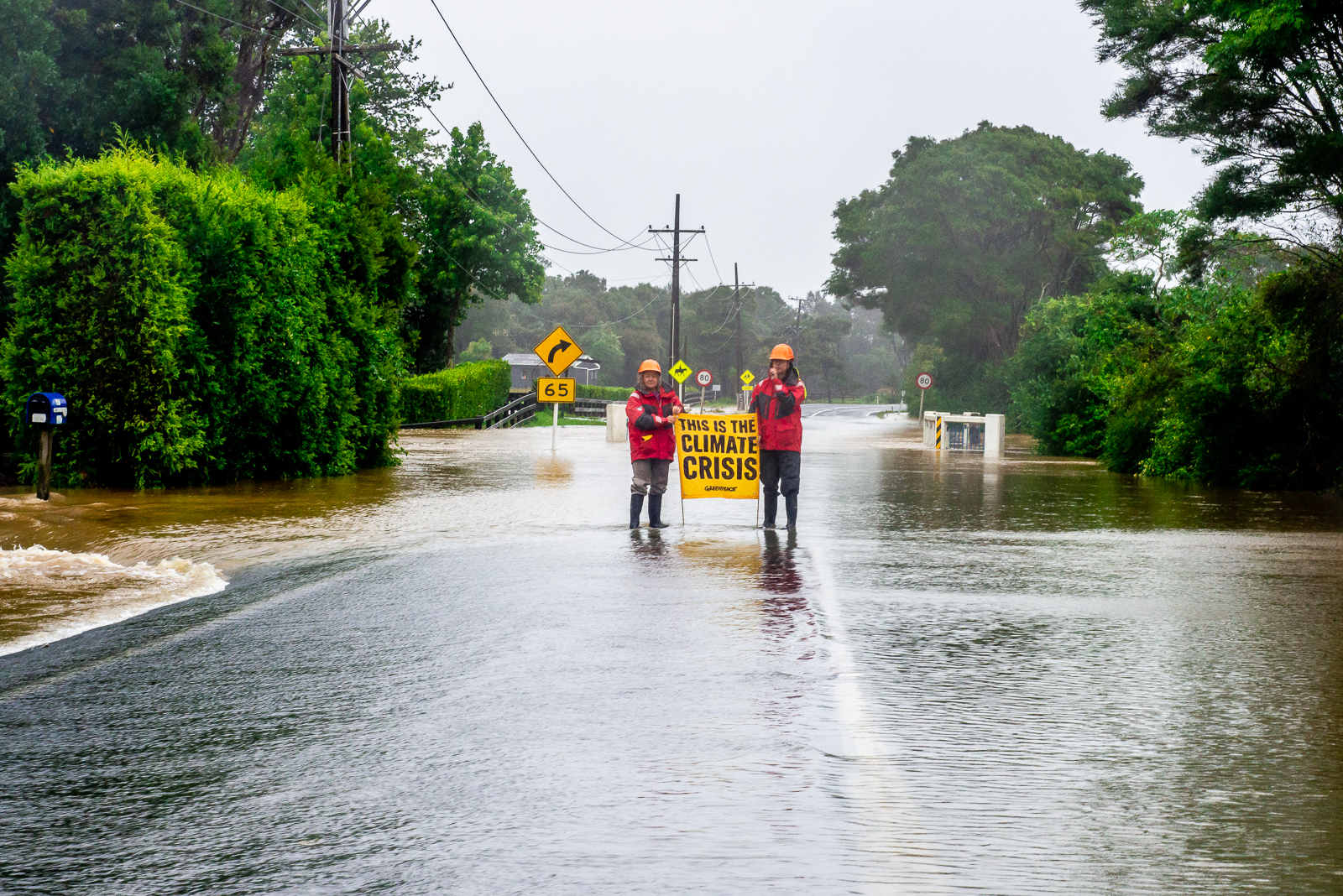 Two people stand in the middle of a road with the surface underwater. The have red jackets and hardhats. They hold a banner saying This is the climate crisis