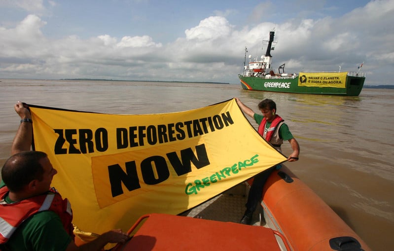 Activists on a dinghy on a brown river water hold a sign saying Zero Deforestation now, with Greenpeace logo. A Greenpeace ship is in the background. 