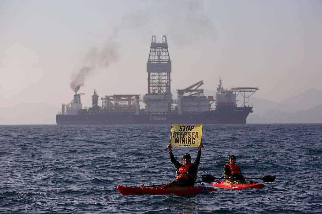 Two Greenpeace activists in kayaks hold a sign saying "Stop Deep Sea Mining". Behind them, is a deep sea mining vessel.