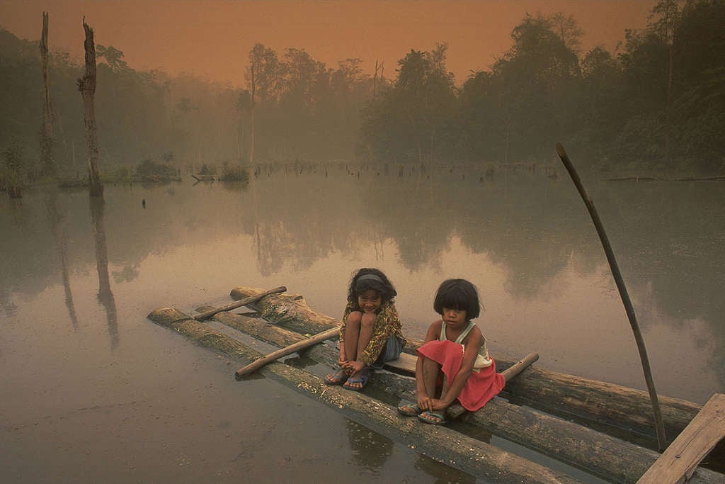 Niños sentados en una balsa de madera en un lago/río rodeado de humo.