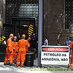 pessoas simulam a exploração de petróleo com uma máquina e barris em frente ao prédio do governo.