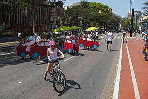 No primeiro plano, pessoas vestidas de carros promovem um pequeno "congestionamento" e impedem a passagem de pedestres pela Avenida Paulista; com perspectiva da avenida em dia de sol