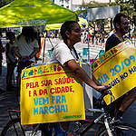 Garota de tranças segura placa com os dizeres "Pela sua saúde e a da cidade, vem pra marcha!"; ao fundo, homem de óculos segura placa com os dizeres "dia 20/09, às 16 horas no Masp"; ambos estão de bicicleta na Avenida Paulista; ao fundo, guarda-sol do Greenpeace
