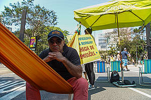 Homem de meia idade sentado na rede no meio da Avenida Paulista. Ao fundo, guarda-sol do Greenpeace e cartaz contra as emissões de gases poluentes
