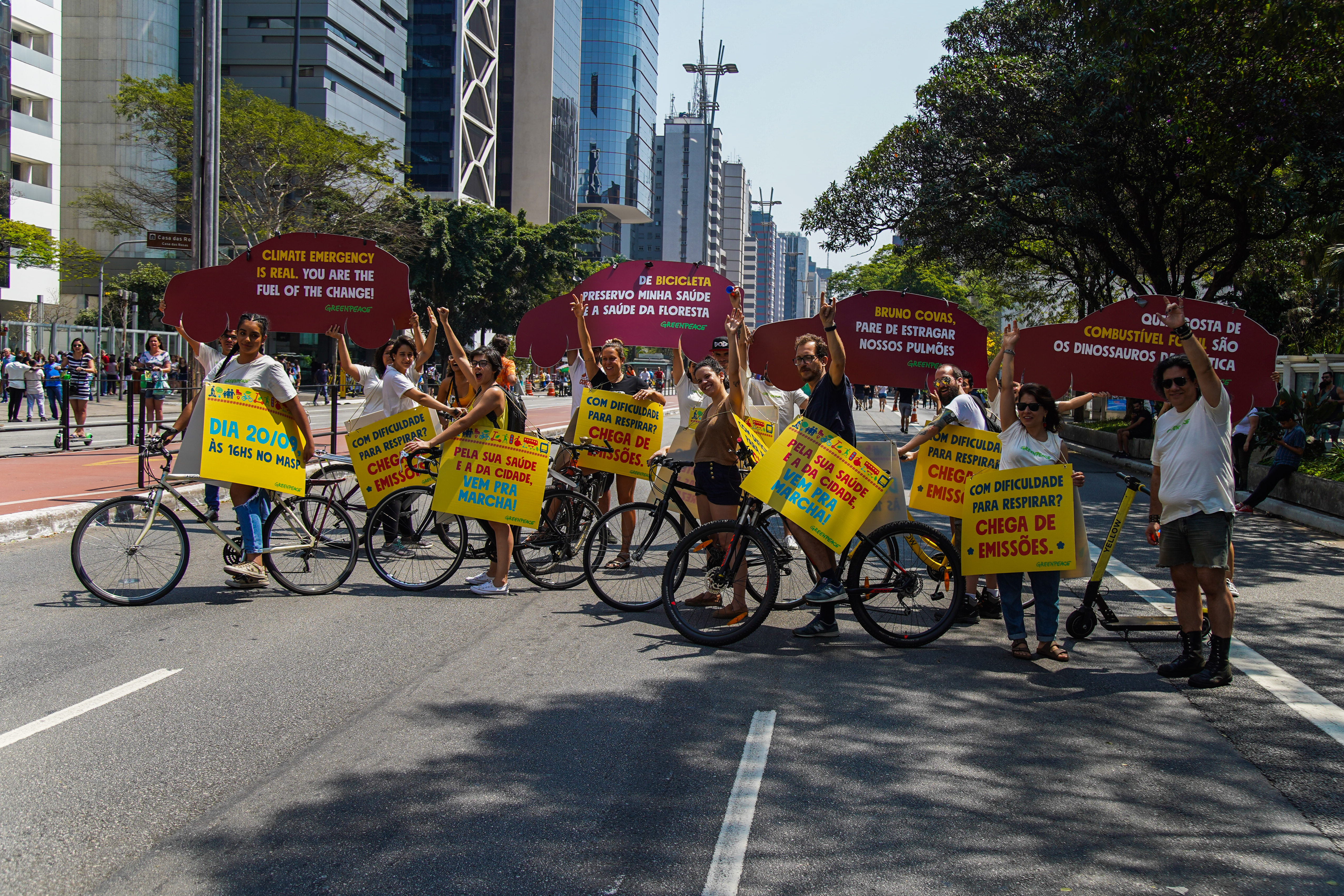 Ativistas do Greenpeace levantam placas com os dizeres "De bicicleta preservo minha saúde e a saúde da floresta", "Bruno Covas, pare de estragar nossos pulmões", Quem gosta de combustível fóssil são os dinossauros da política", com Avenida Paulista em perspectiva, dia de sol