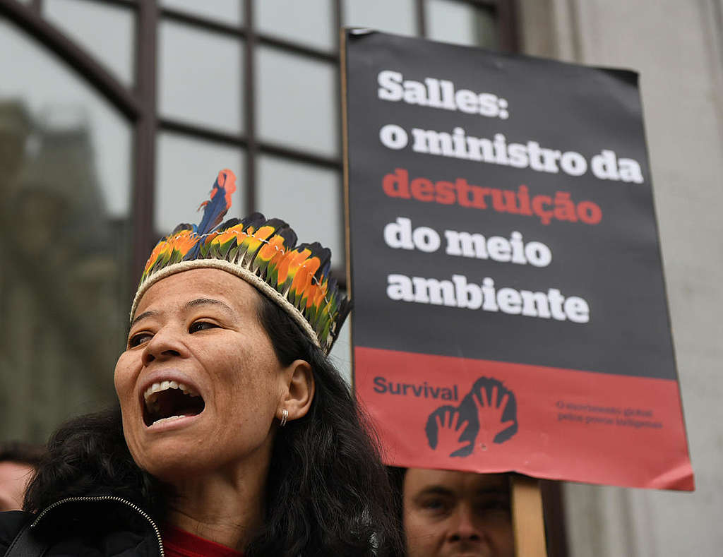 Ativistas protestam em frente à Embaixada do Brasil em Londres, close em mulher usando um cocar e placa ao fundo com a frase "Salles: o ministro da destruição do meio ambiente"