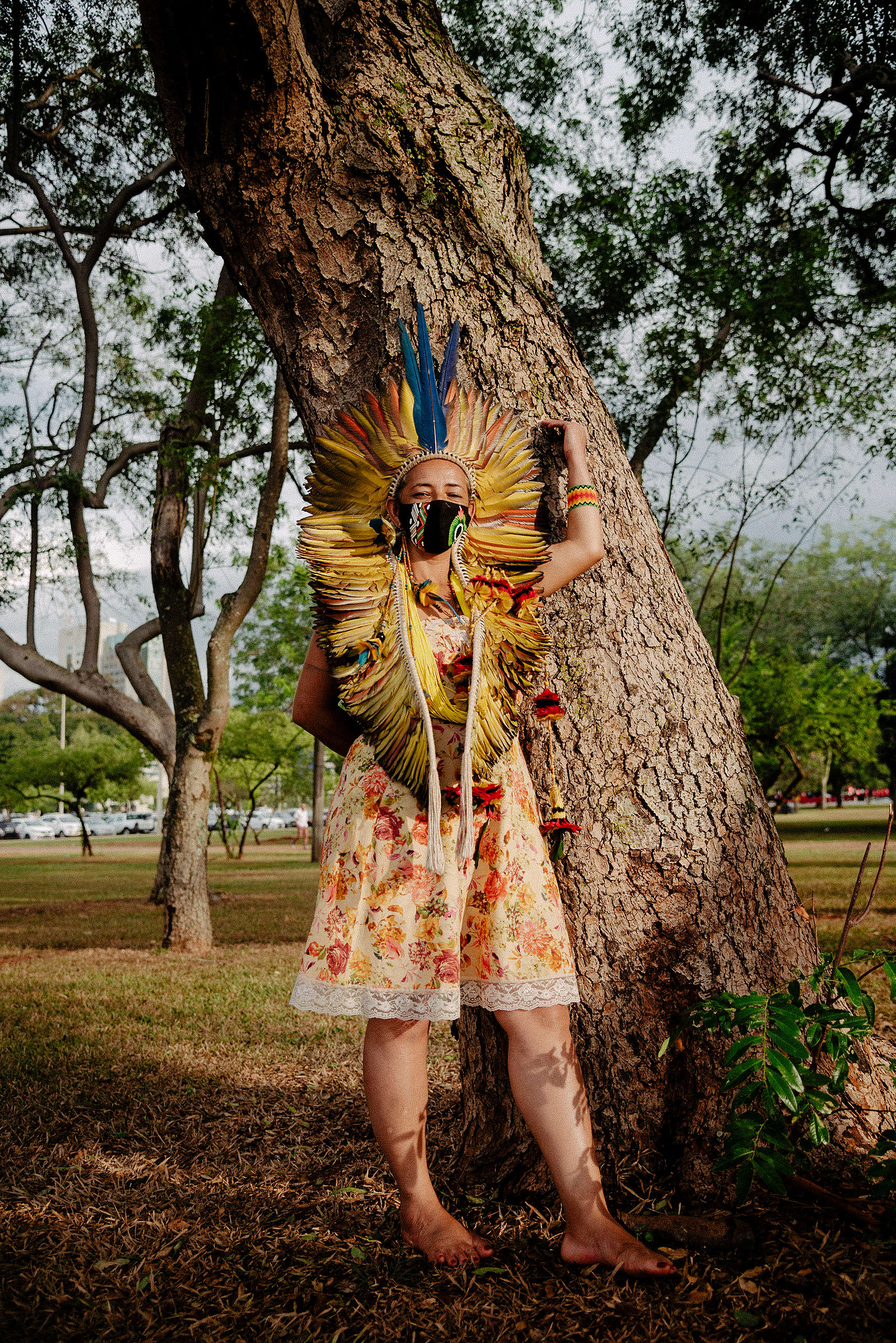 Indígena mulher, com roupa colorida e cocar com penas amarelas, vermelhas e azuis enconstada em uma árvore. Ao fundo, paisagem formada por árvores, grama e carros estacionados.