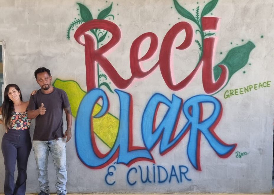 Foto de duas pessoas voluntárias ao lado de uma parede com o grafite escrito: "Reciclar é cuidar - Greenpeace"