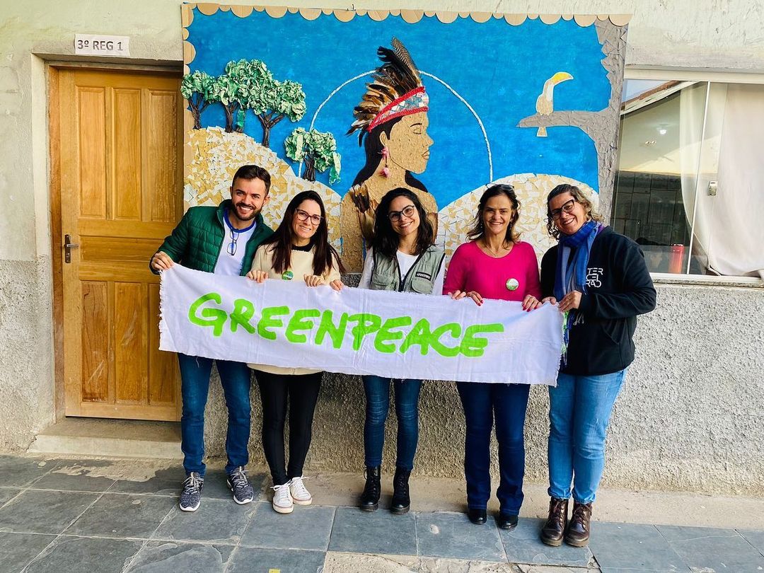 Foto de pessoas voluntárias em ambiente externo, eles e elas seguram o banner escrito Greenpeace