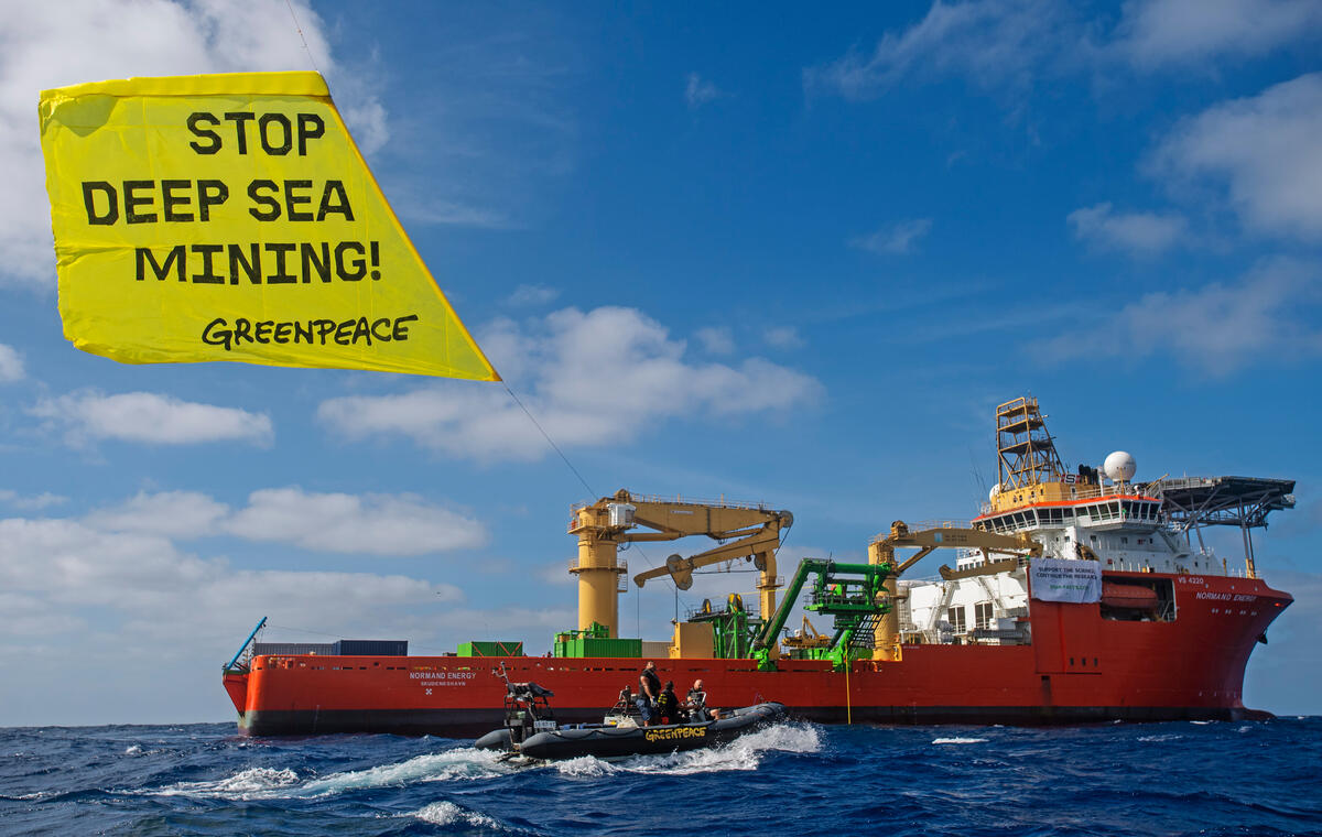 Um navio responsável pela mineração em águas profundas em alto-mar, ao lado de um bote hastando uma bandeira amarela com os dizeres "Stop Deep Sea Mining", que quer dizer "Parem a mineração em águas profundas", em inglês