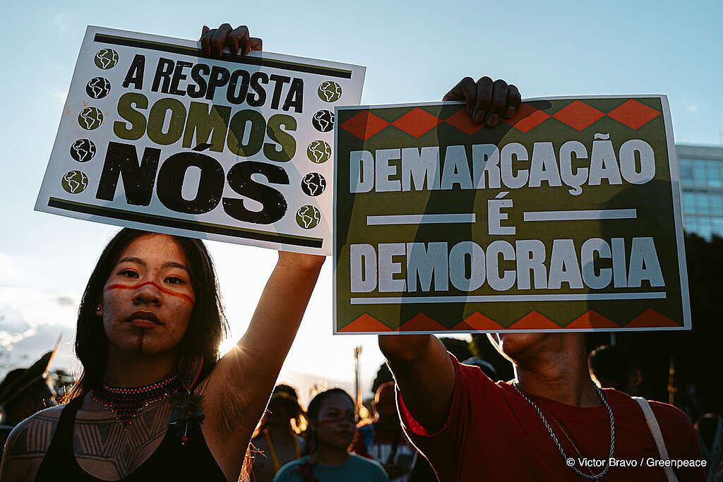 Duas mulheres indígenas com semblante sério segurando duas placas escritas: "A resposta somos nós" e "Demarcação é Democracia". Elas participaram da Marcha no Acampamento Terra Livre, em Brasília