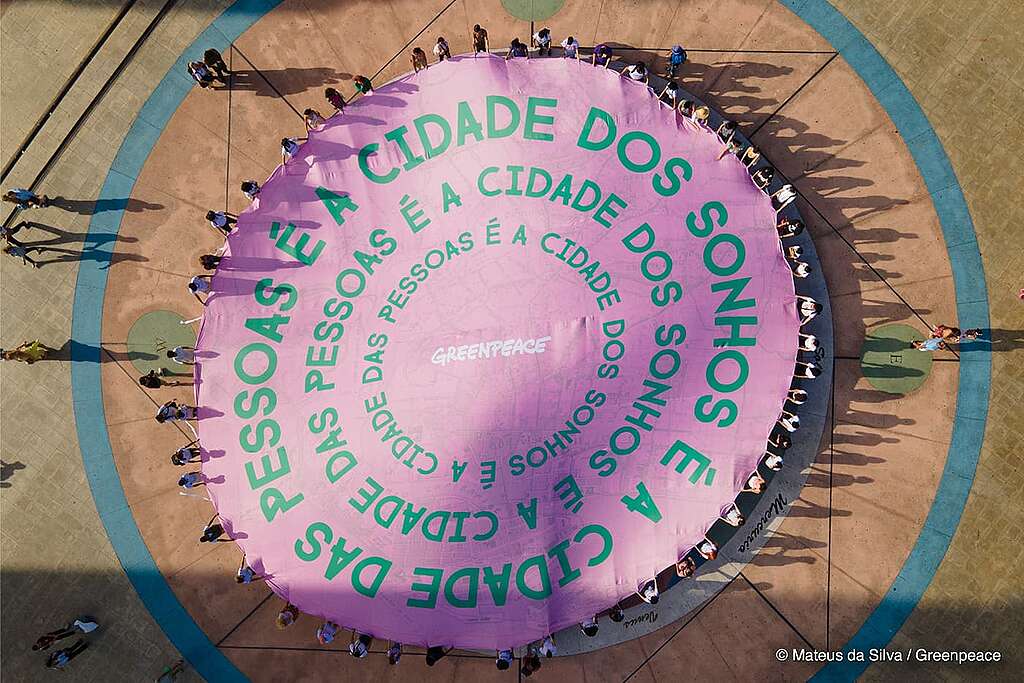 Foto tirada de cima com cerca de 100 pessoas de diferentes etnias segurando um banner redondo escrito no centro "Cidade dos sonhos é a cidade das pessoas".