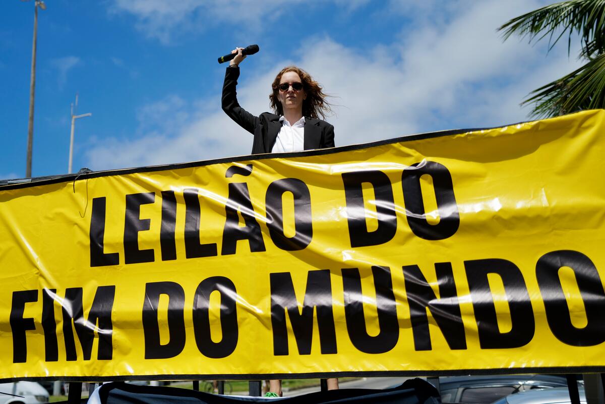  mulher branca com um microfone na mão e semblante sério posicionada em cima de um caminhão com um banner amarelo aberto em sua frente. No banner tem escrito: Leilão do fim do mundo.