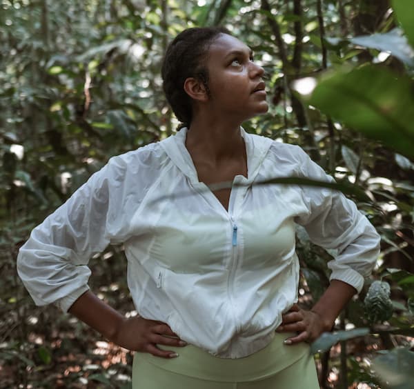 mulher negra com semblante sério posicionada em área preservada na Amazônia com os braços na cintura.