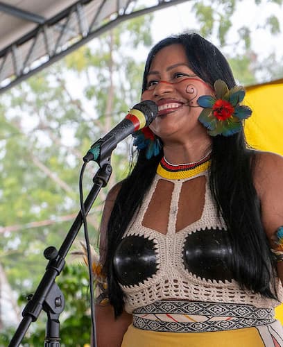 mulher indígena de cabelos longos com semblante sorridente posicionada em um palco com um microfone à sua frente.