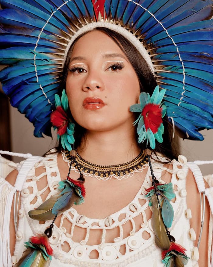 mulher indígena com semblante sério e cabelos castanhos veste um cocar na cabeça.