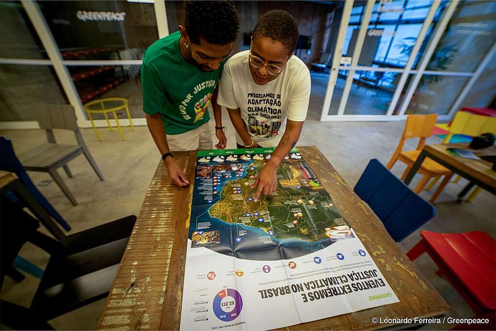 homem e mulher em pé posicionados em frente a uma mesa de madeira, na mesa tem o mapa "Por que lutar por justiça climática" aberto e ambos estão lendo o conteúdo do mapa. 