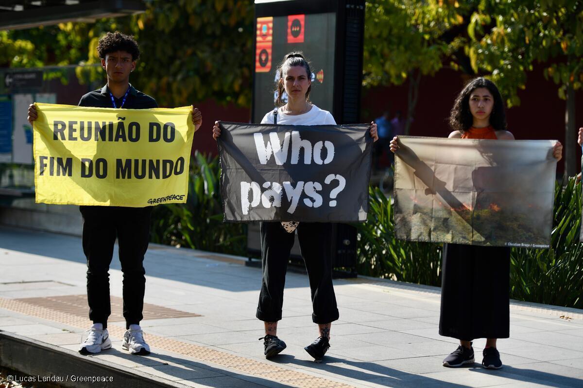 Três ativistas protestam com cartazes em frente a uma das maiores conferências de petróleo e gás do mundo, sediada no Rio de Janeiro, contra a exploração de petróleo no Brasil. O primeiro cartaz tem os dizeres: Reunião do fim do mundo, o segundo: "Who pays? - Quem paga, em português, e o terceiro uma imagem das queimadas que vem desvastando o país.