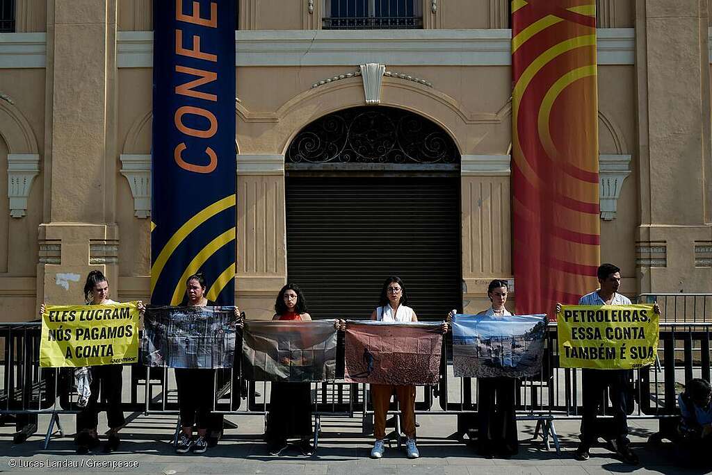 Seis ativistas estão com cartazes  em frente a uma das maiores conferências de petróleo e gás do mundo, sediada no Rio de Janeiro, contra a exploração de petróleo no Brasil. O primeiro cartaz tem os dizeres: Eles lucram, nós pagamos a conta, seguido de cartazes com imagens da devastação que a crise climática está causando em nosso país, e o último cartaz com os dizeres: petroleiras, essa conta também é sua.