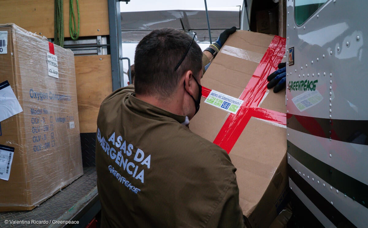 Imagem mostra pessoa de costas carregando caixa de equipamentos para o avião do Greenpeace, que levará suprimentos de saúde para o Alto Rio Negro. No macacão da pessoa lê-se: Asas da Emergência, Greenpeace.