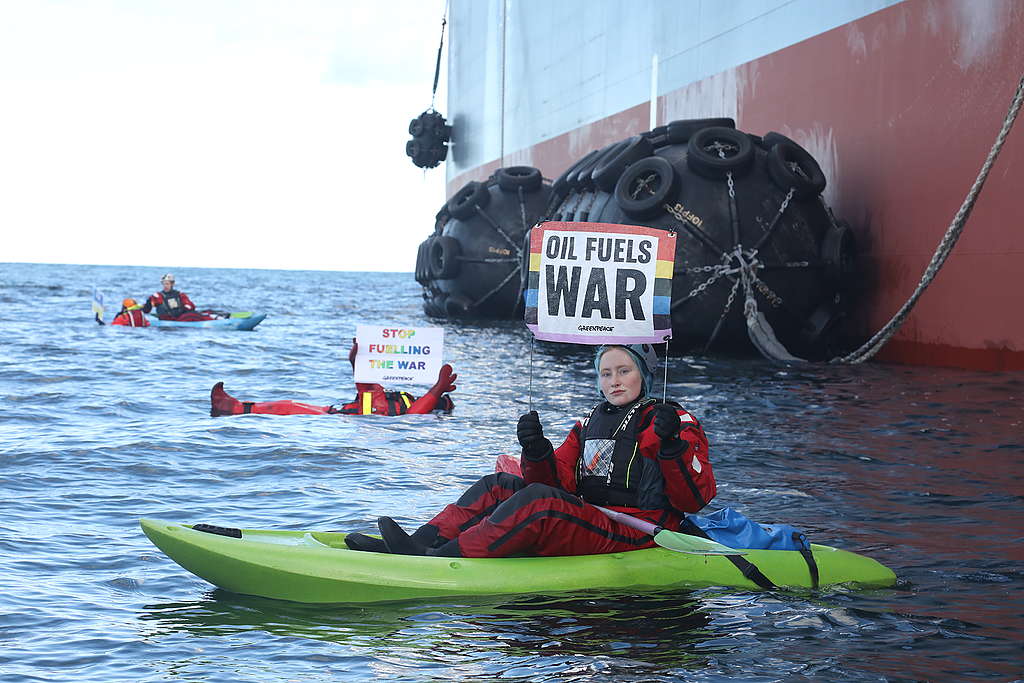Greenpeacen aktivisteja kellumassa sekä kajakkien päällä meressä Venäläisen öljytankkerin vieressä. Aktivisteillä on kylttejä, joissa lukee "oil fuels war" ja "stop fuelling the war".