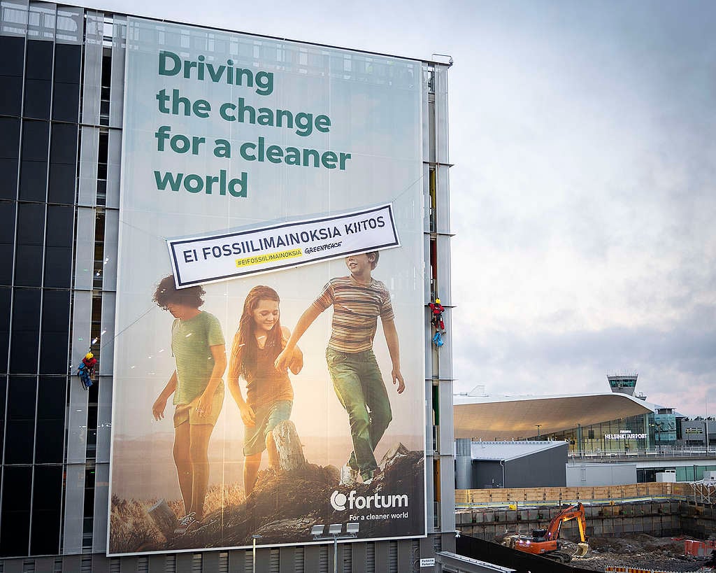 Fortumin mainos Helsinki-Vantaan lentoasemalla, jossa lukee "Driving the change for a cleaner world". Mainoksen päällä on Greenpeacen banderolli tekstillä "Ei fossiilimainoksia, kiitos".
