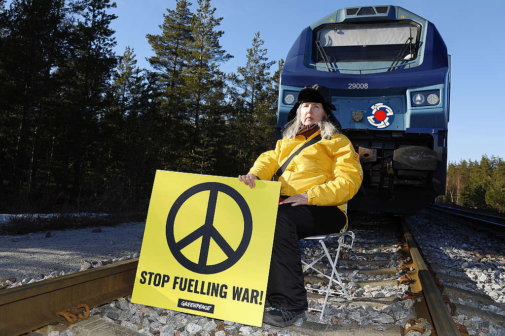 Greenpeacen vapaaehtoinen istuu raiteilla estäen venäläisen hiilijunan liikkumisen. Vapaaehtoisella on kädessä kyltti, jossa rauhanmerkki, ja jossa lukee "stop fueling war".