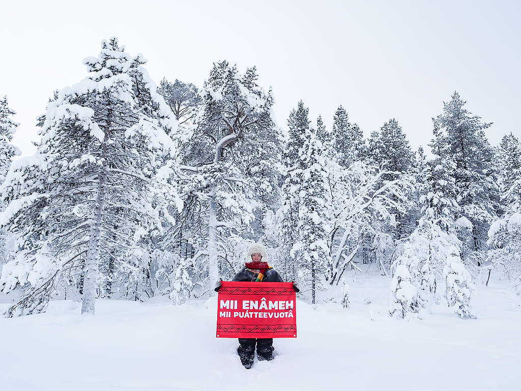 Kuvassa saamelainen poronkasvattaja pitelee banneria, jossa lukee Inarinsaameksi "Meidän maa, meidän tulevaisuus". Taustalla on talvista metsämaisemaa.