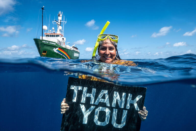 Aktivisti kellumassa meressä pitäen kädessä kylttiä, jossa lukee "THANK YOU". Takana Greenpeacen Arctic Sunrise -laiva.