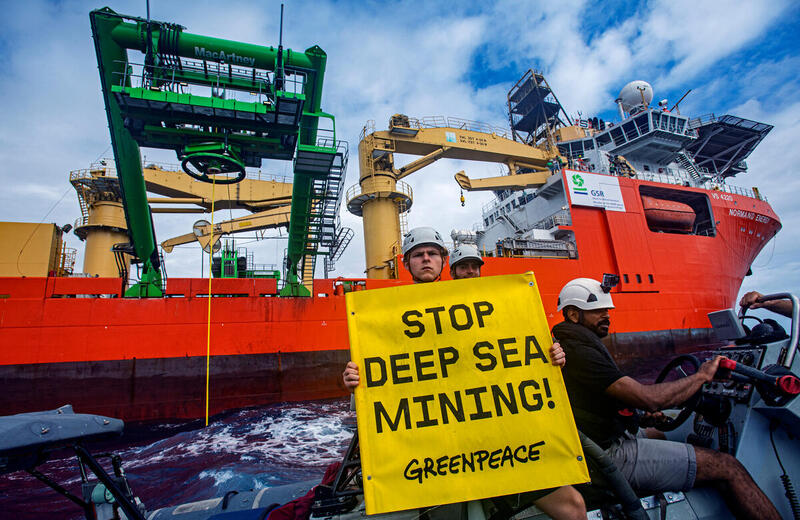 Kolme Greenpeacen aktivistia ison kaivoslaivan edessä. Yhdellä aktivisteista on kädessä kyltti,  jossa lukee "STOP DEEP SEA MINING".