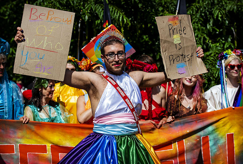 Picture showing a Queers4Climate activists standing against climate breakdown and holding two signs reading "Be proud of who you are" and "Pride is Protest".