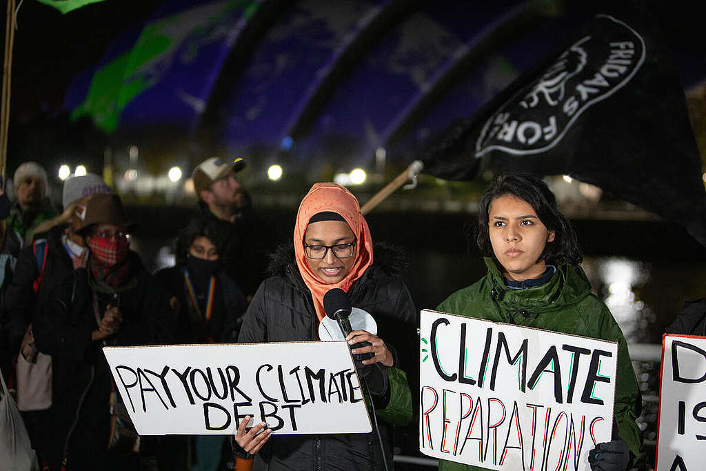 A girl with a head scarf speaking into a mic while holding a placard that says Pay Your Cimate Debt. Another girl is standing next to her holding a placard that says Climate Reparations.