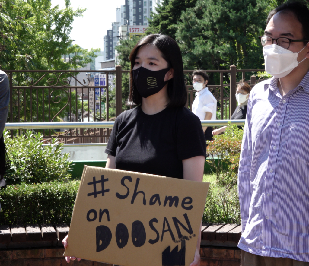 A girl with shoulder length hair holding a sign that has the text #Shame on Doosan on it. There is a man standing next to her.