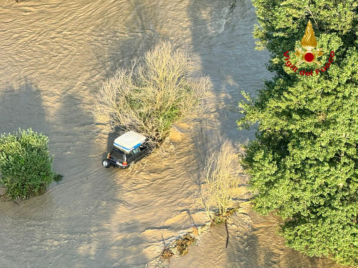 A partially submerged car is pinned against a tree in a flooded open area.