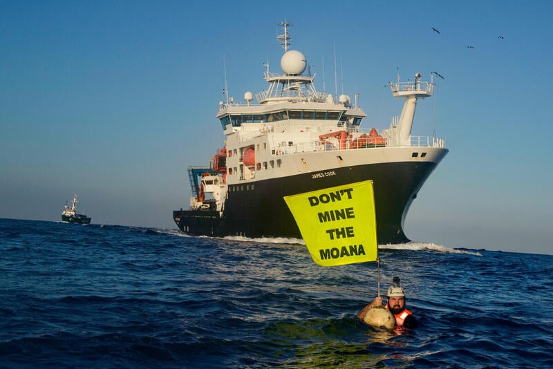 Aktivist foran skipet James Cook i Stillehavet, med et flagg med teksten “Don't Mine the Moana" (Ikke anlegg gruver i havet).