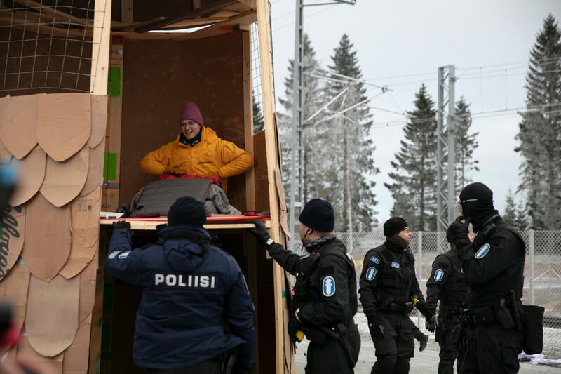 En glad aktivist från Greenpeace sitter inuti en stor uggla av pappkartong när polisen får tag på honom. Vinterlandskap i bakgrunden. 